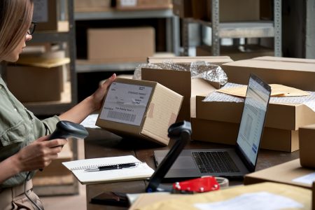 Female seller worker online store holding scanner scanning parcel barcode tag packing ecommerce post shipping box checking online retail store orders in dropshipping delivery service warehouse.